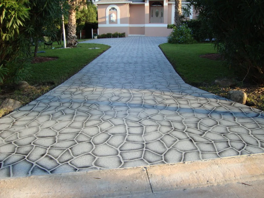 Concrete walkway with a decorative stone-like pattern leading to a house, flanked by green grass and bushes.