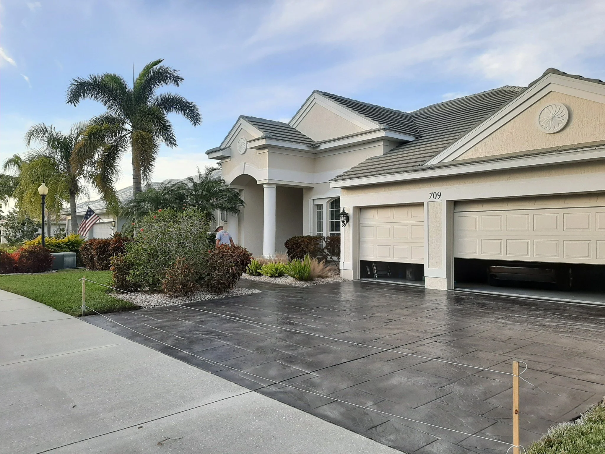 Modern house with a two-car garage, landscaped front yard with palm trees and shrubs, and a driveway with recently sealed black asphalt.