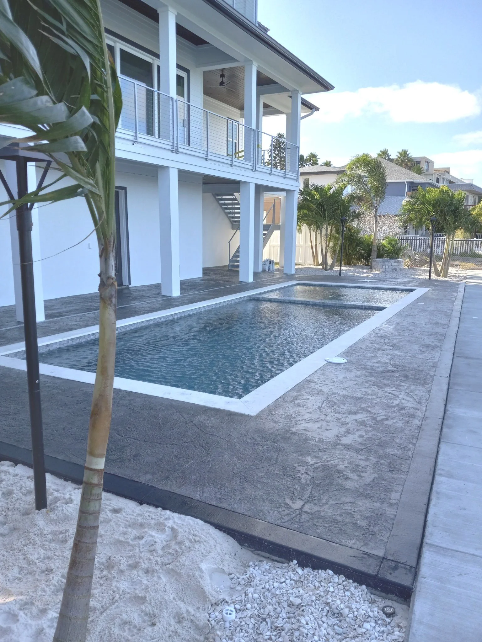Outdoor backyard with a small rectangular swimming pool, a upper balcony with metal railing, surrounding trees, and neighboring houses in the background.