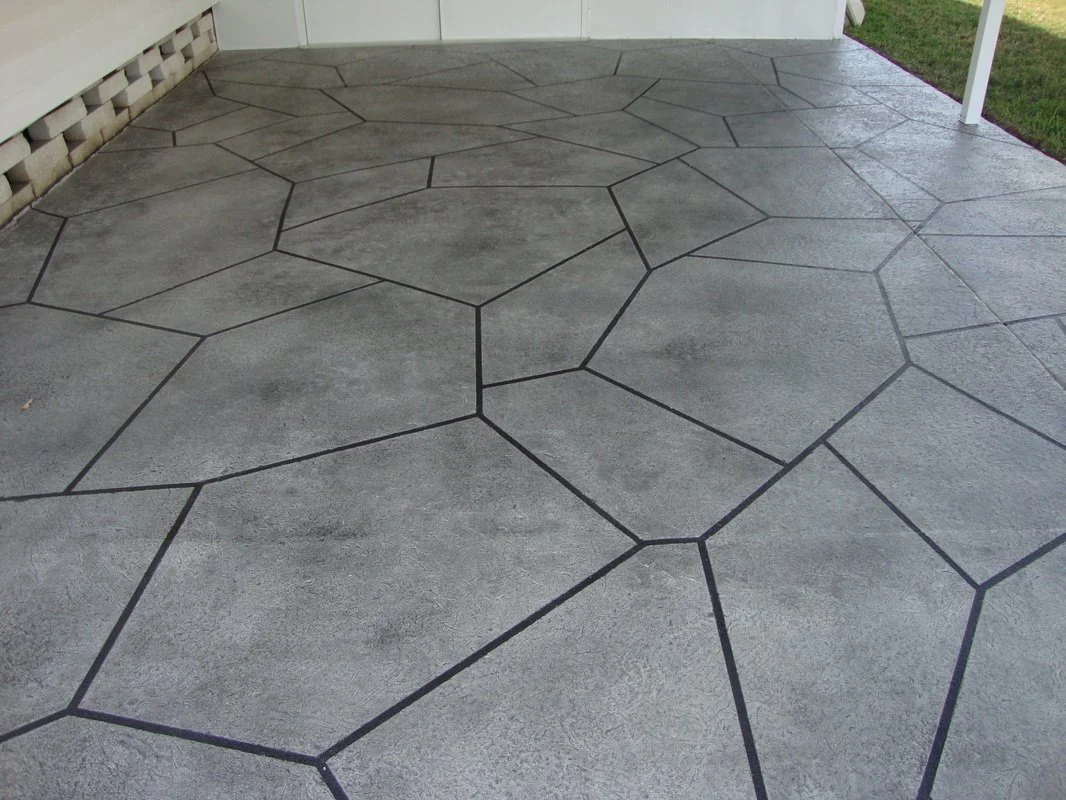 Concrete patio with interlocking irregular hexagon pattern and black grout lines, adjacent to a white house and green lawn.