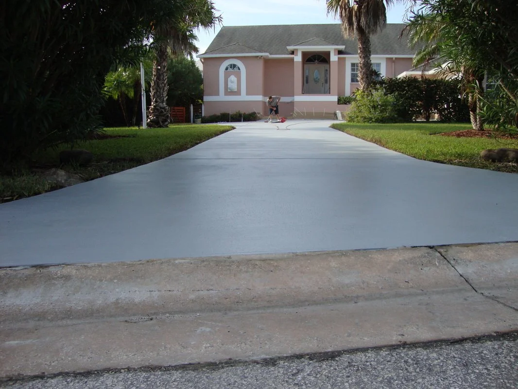 Freshly paved light gray driveway leading to a pink house with white trim, front yard with green grass and tropical bushes, with two palm trees on either side of the house, in a suburban neighborhood.