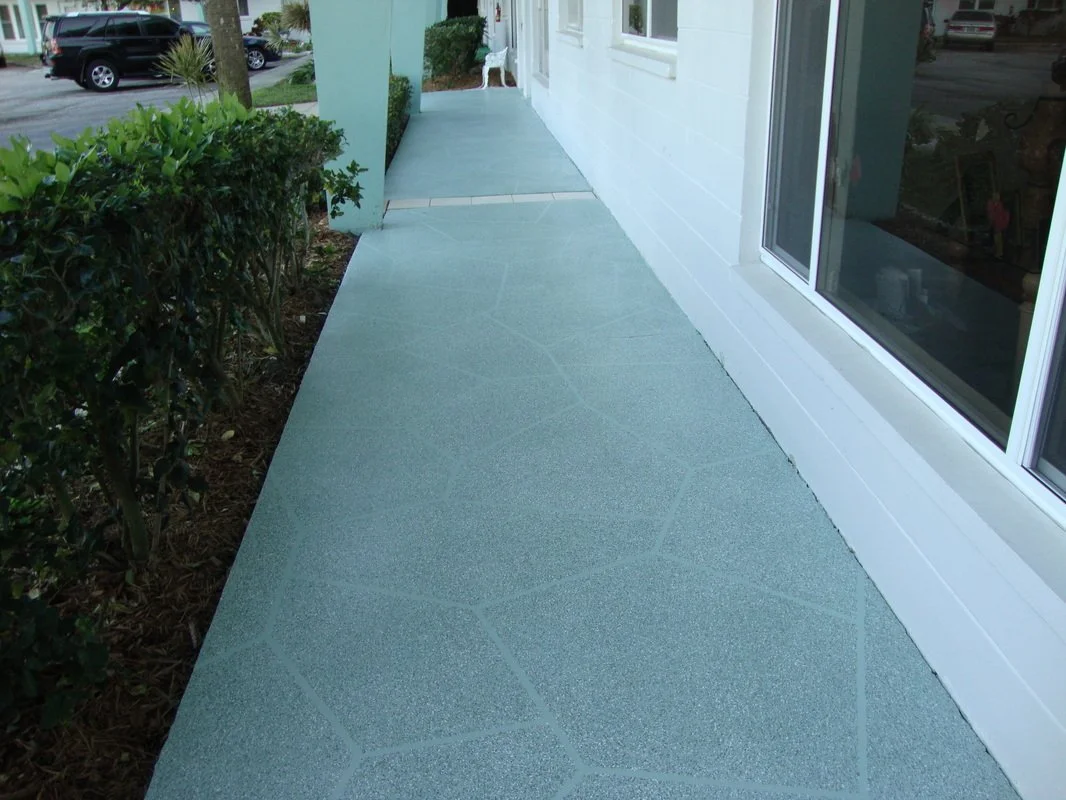 View of a concrete sidewalk next to a white house with large windows and a garden bed with bushes on the left, in a residential neighborhood.