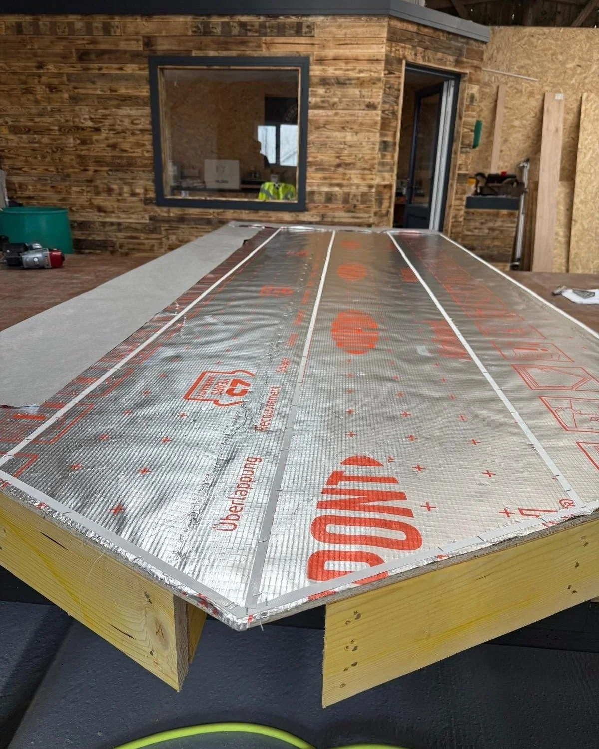 Construction workspace with insulated material on a wooden table, wooden walls, an office in the background.