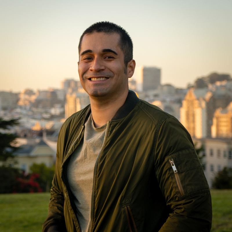 Jason Alarcón smiling in a green jacket standing outdoors with a cityscape background at sunset.