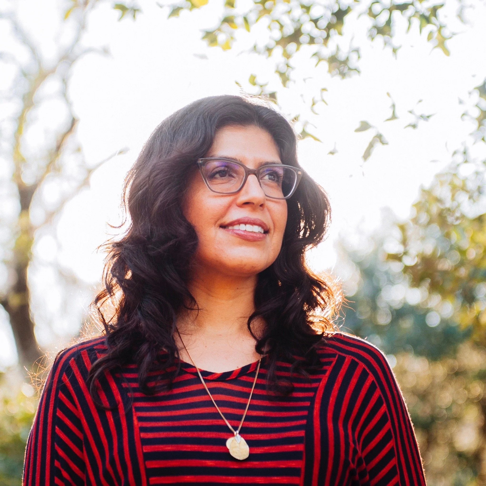 Amy Martinez headshot with long dark wavy hair, glasses, and a striped red and black top standing outdoors with trees and sunlight in the background.