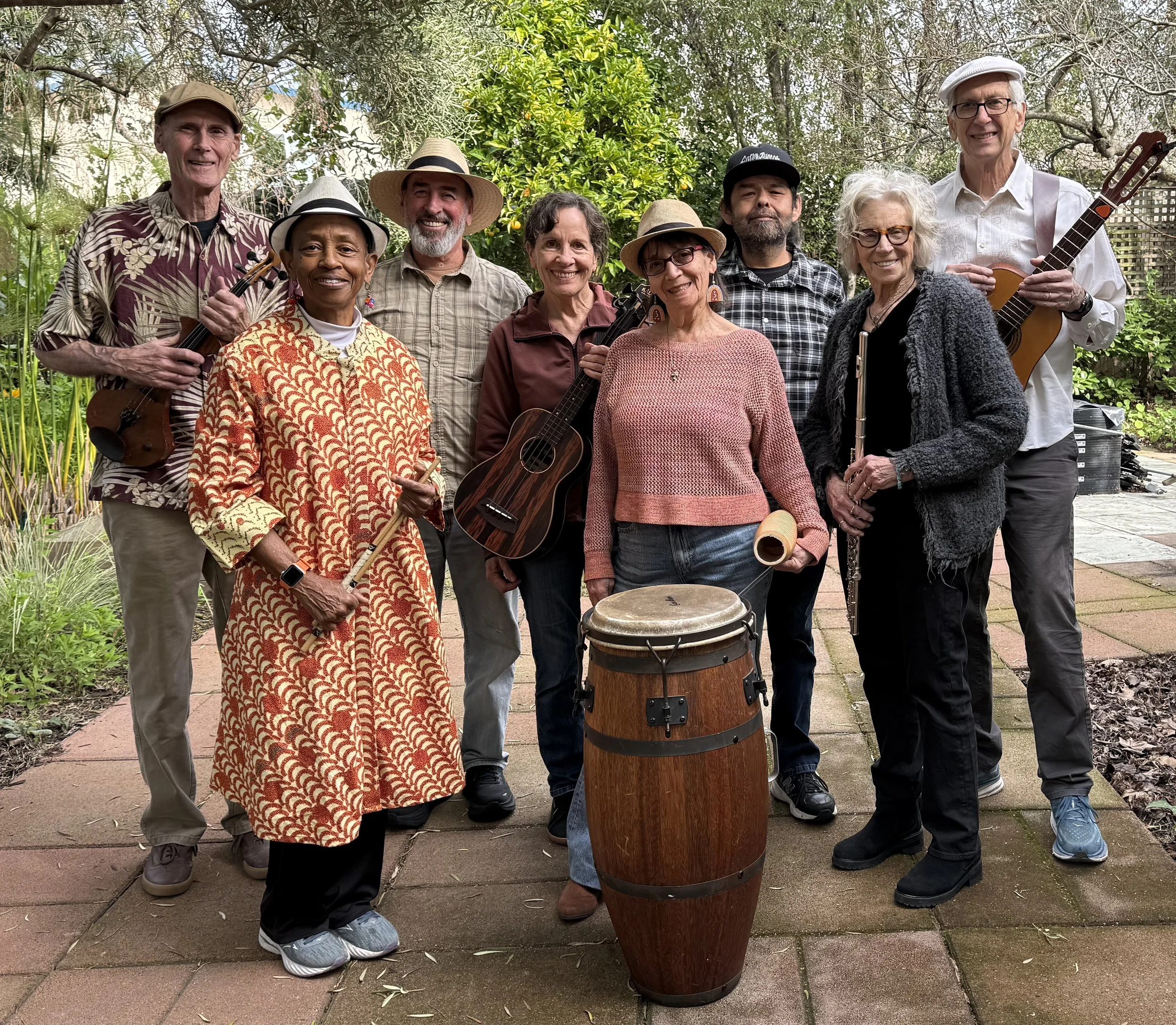 Group of nine salsa musicians outdoors with musical instruments, standing on a garden patio surrounded by green trees and plants.