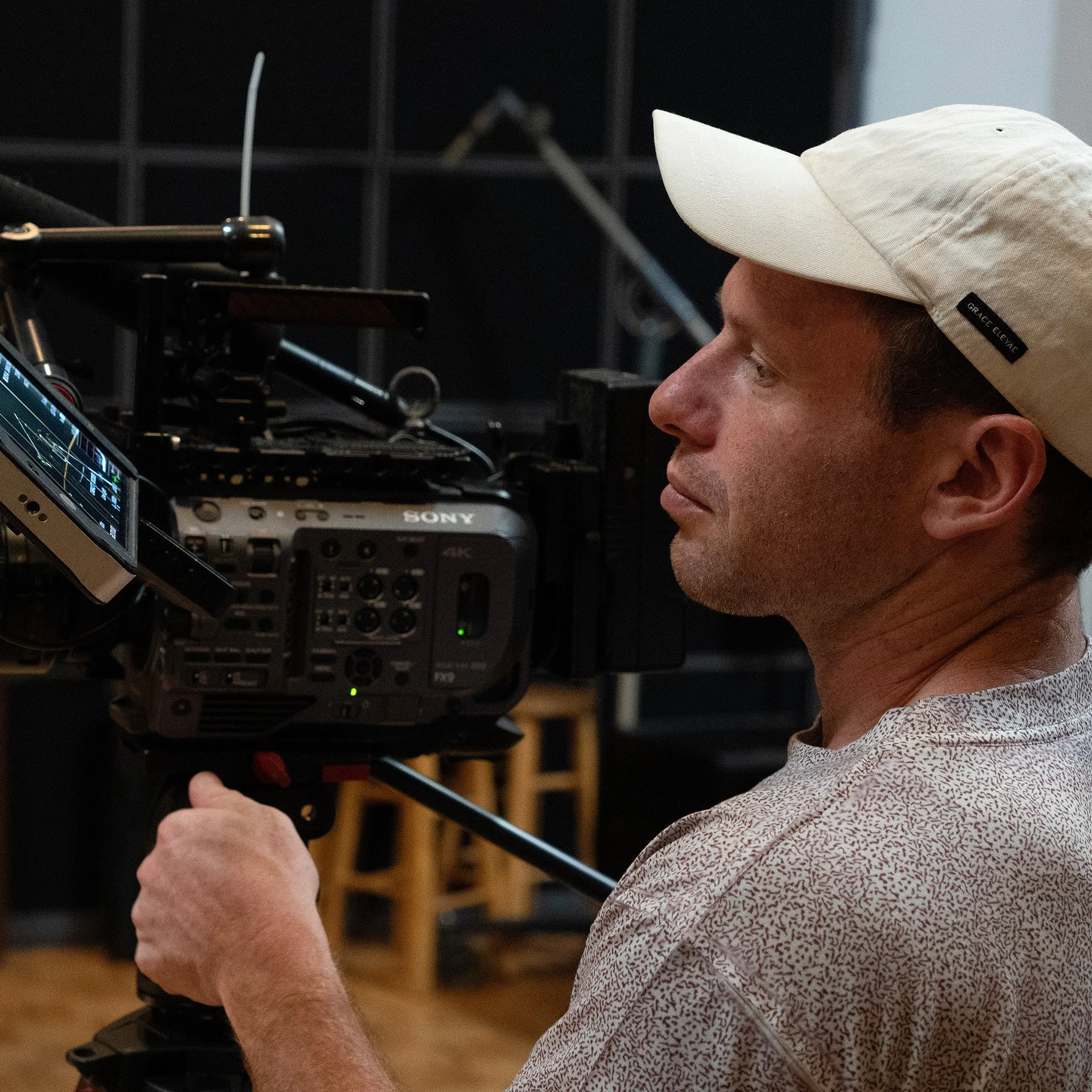 Ethan Goldwater wearing a beige cap and patterned shirt operating a professional video camera while filming the Renegade Queens Documentary.