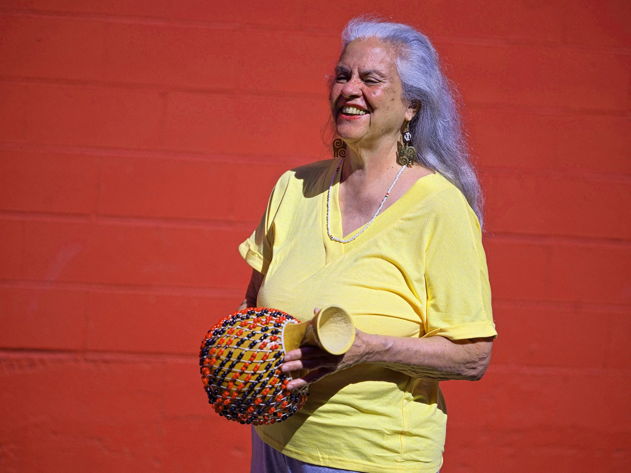 An woman with gray hair smiling while holding a shekere, wearing a yellow t-shirt and jewelry against an orange background.