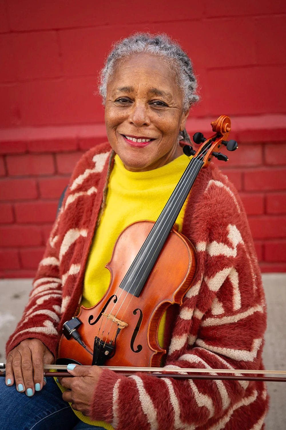 Smiling woman with gray hair holding a violin, wearing a yellow top and a red and white patterned cardigan, standing against a red brick wall.