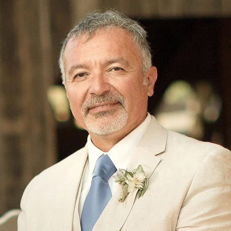 Manuel Tsingaris has a beard, is wearing a cream-colored suit, light blue tie, and a floral boutonniere, and is standing outdoors with a blurred background.