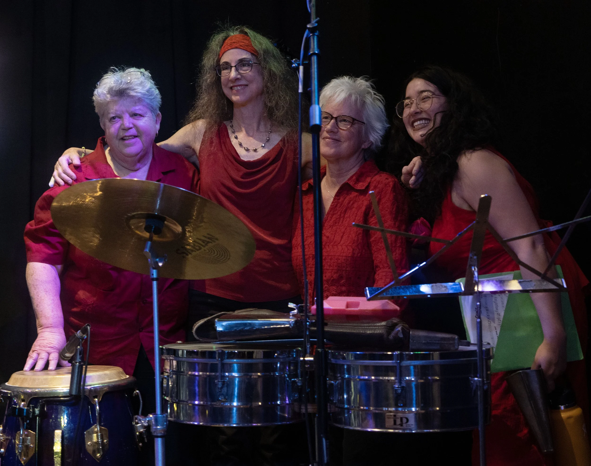 Four women on stage, dressed in red, smiling with musical instruments including timbales and a cymbal, posing after a performance.