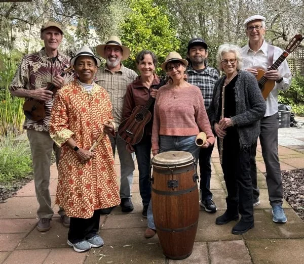 Group of eight people standing outdoors on a paved path, holding musical instruments, with greenery in the background.