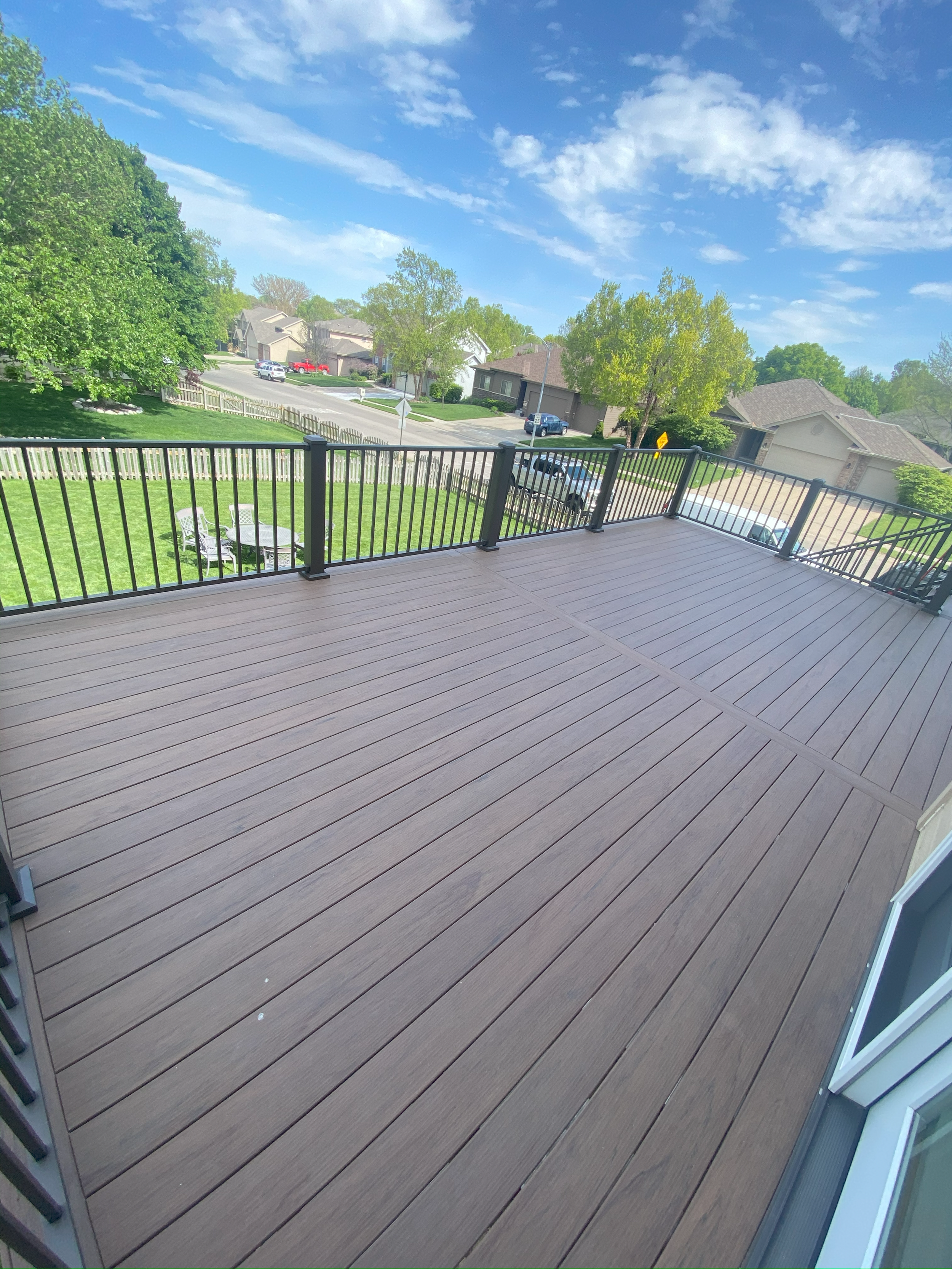 Raised wooden deck with railing overlooking a suburban neighborhood with green lawns, trees, and houses under a blue sky.