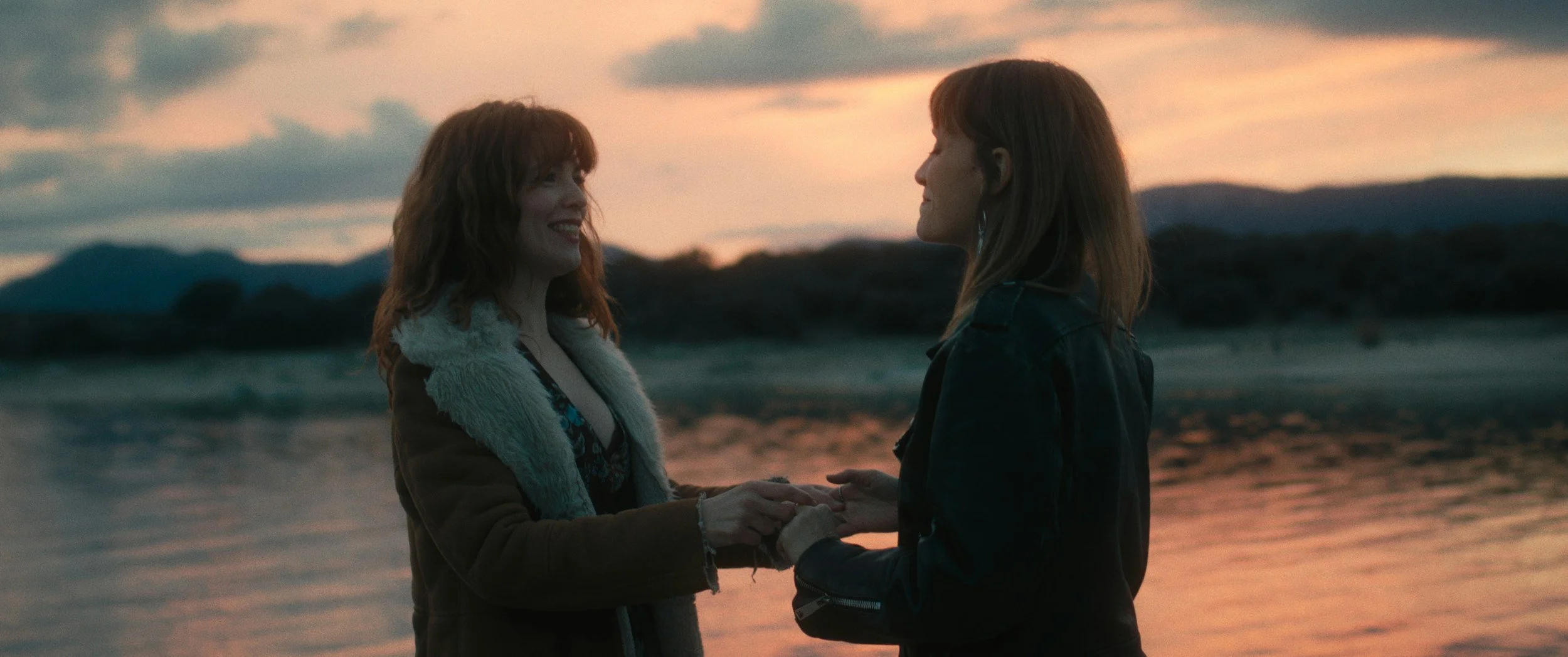 Two women holding hands and smiling at each other on the beach at sunset.
