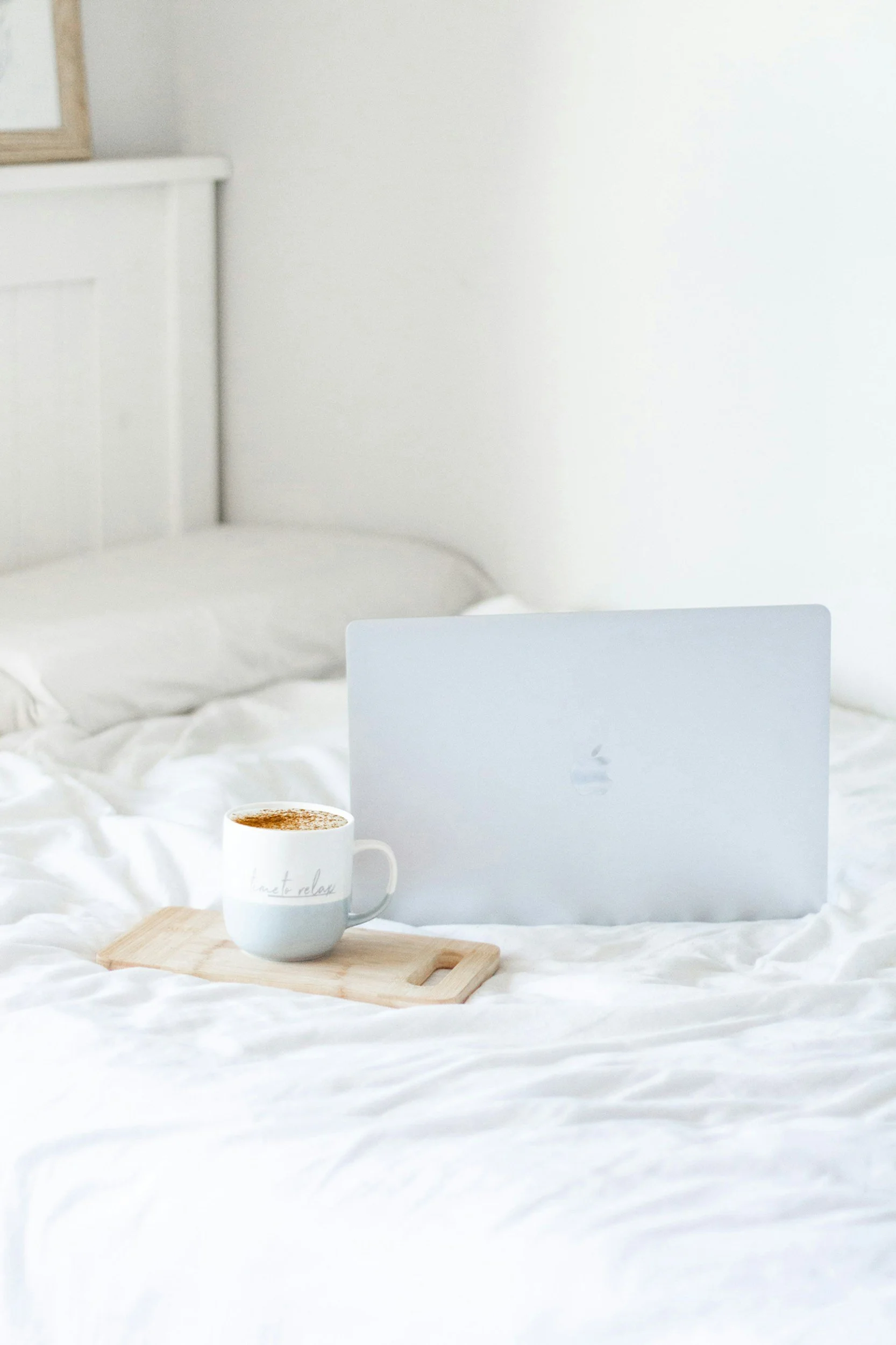 A coffee mug with the words "time to relax" written on it, placed on a wooden tray on a white bedspread. In the background, a silver laptop and a white wall are visible.