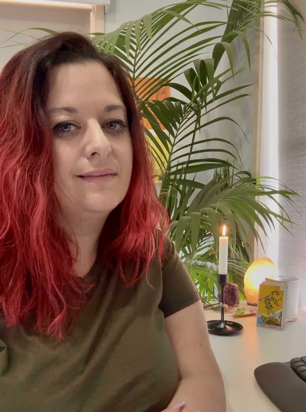 A woman with red hair sitting at a table with a spiritual or meditative setup, including a candle, salt lamp, tarot card, and crystals, with green plants in the background.