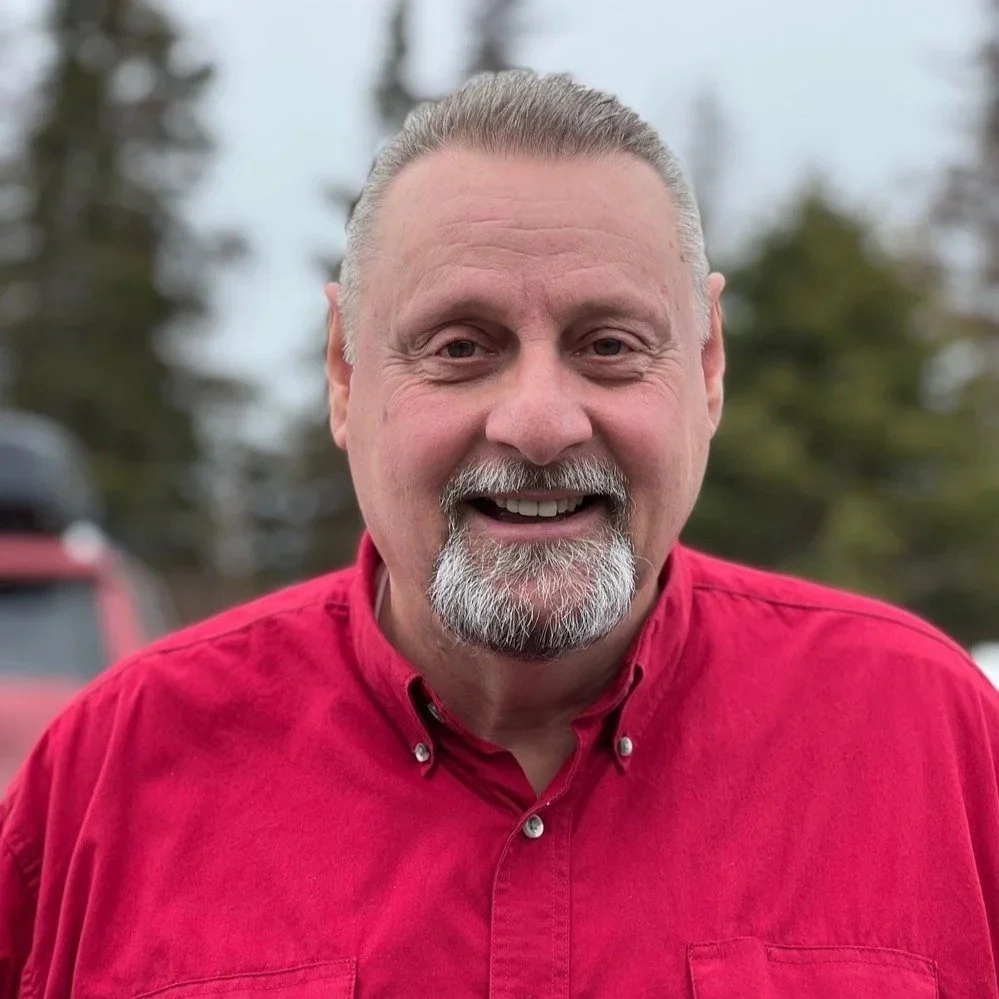 A smiling middle-aged man with gray hair and a beard, wearing a red button-up shirt, outdoors with blurred trees and vehicles in the background.