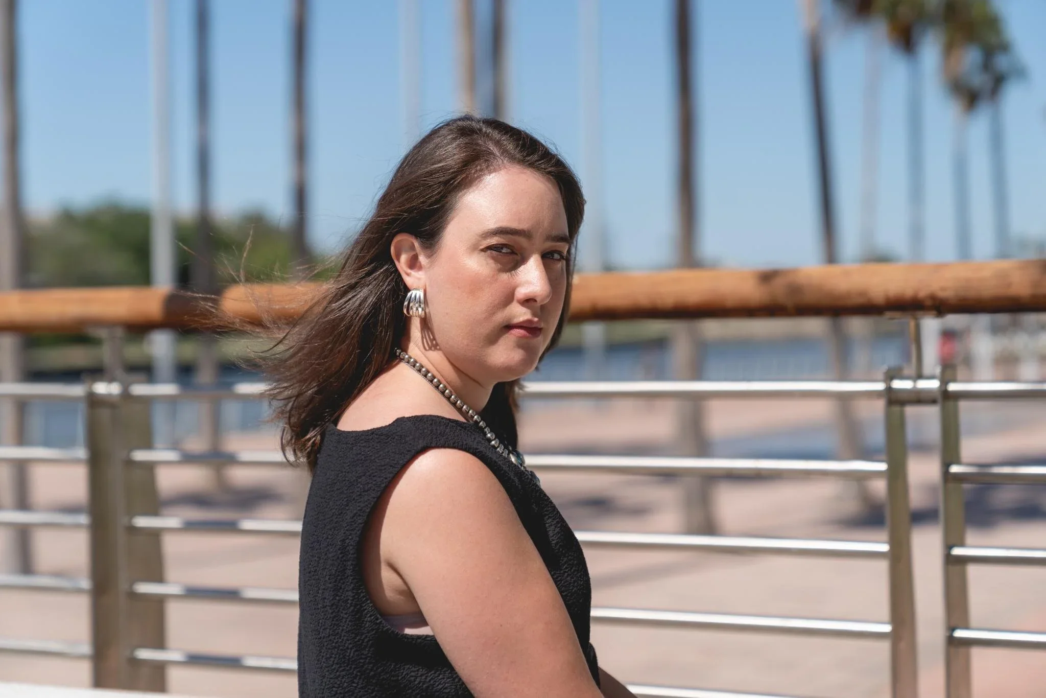 A woman with shoulder-length brown hair styled in a bob, wearing a black sleeveless top, silver earrings, and a pearl necklace, standing outdoors near a metal railing with a waterfront background and palm trees.