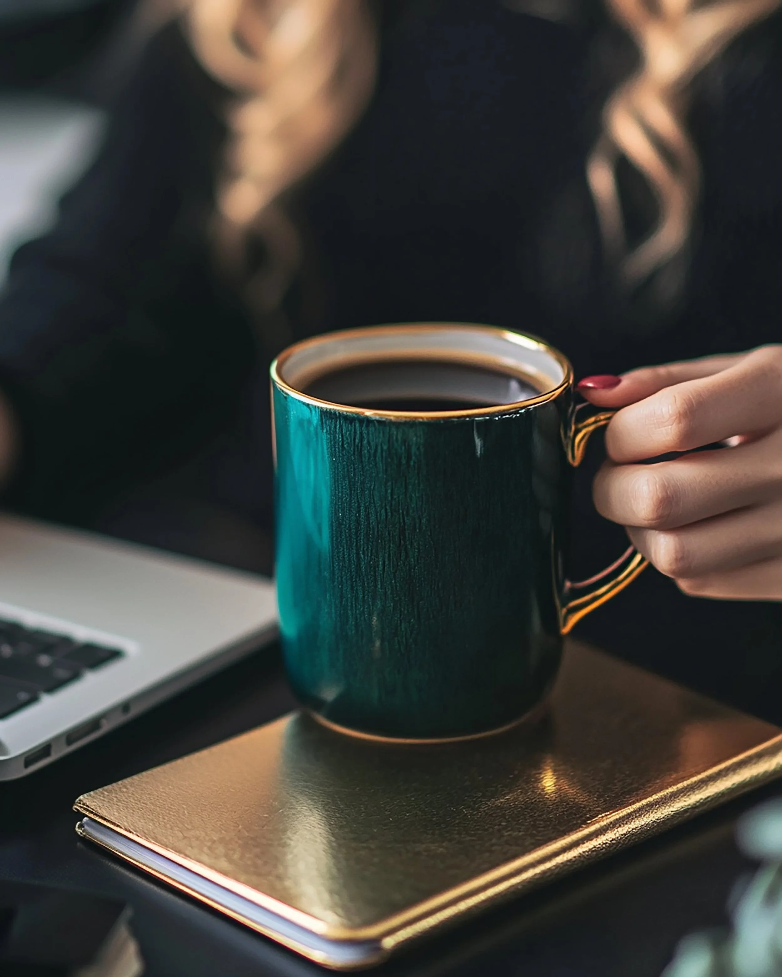 A person's hand holding a dark green ceramic mug with gold trim, filled with coffee, on a gold-colored notebook, with a laptop partially visible in the background.