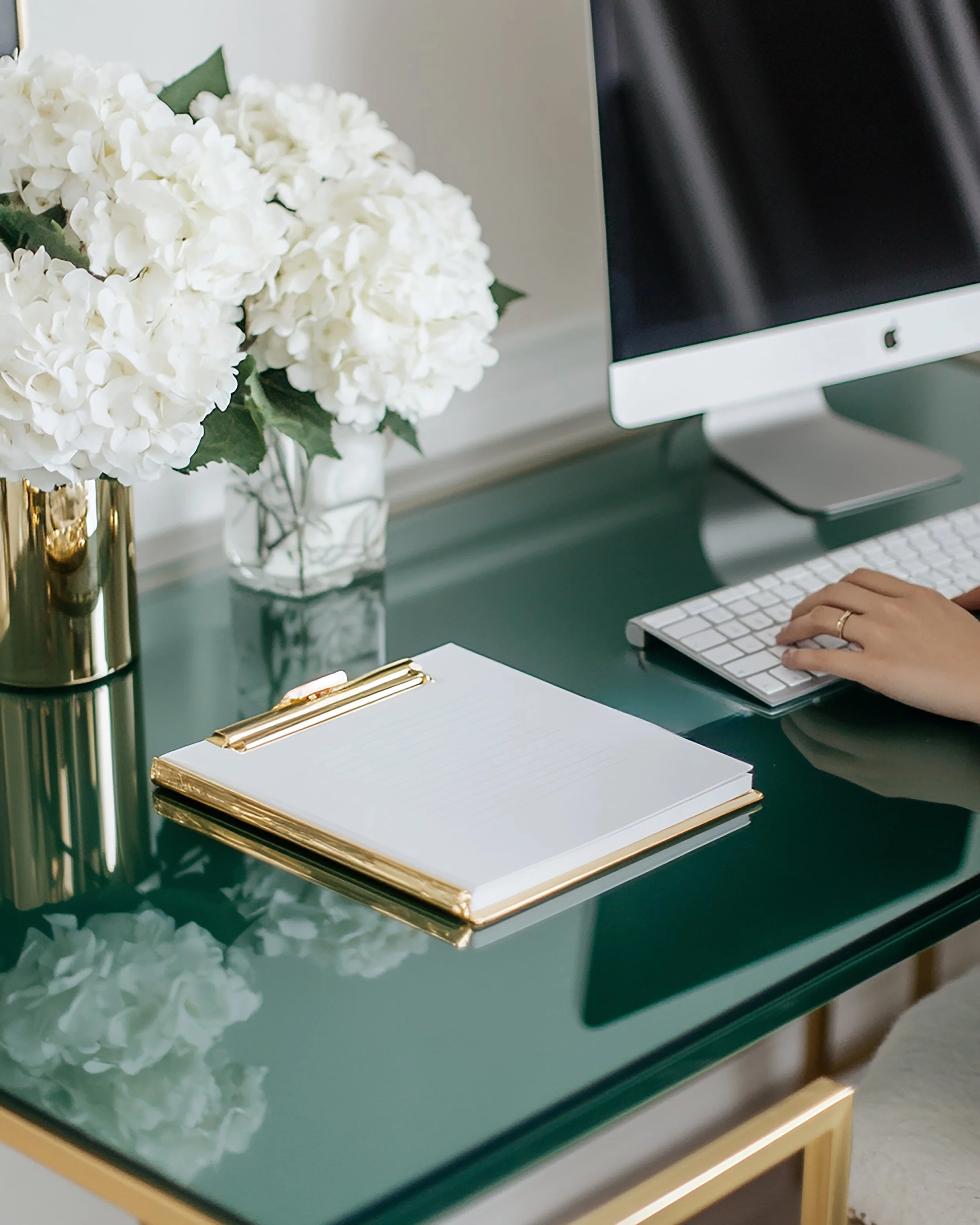 A green glass desk with gold trim, featuring a white notepad with gold binder clip, a vase with white hydrangeas, an iMac computer, and a person's hand typing on a white keyboard.