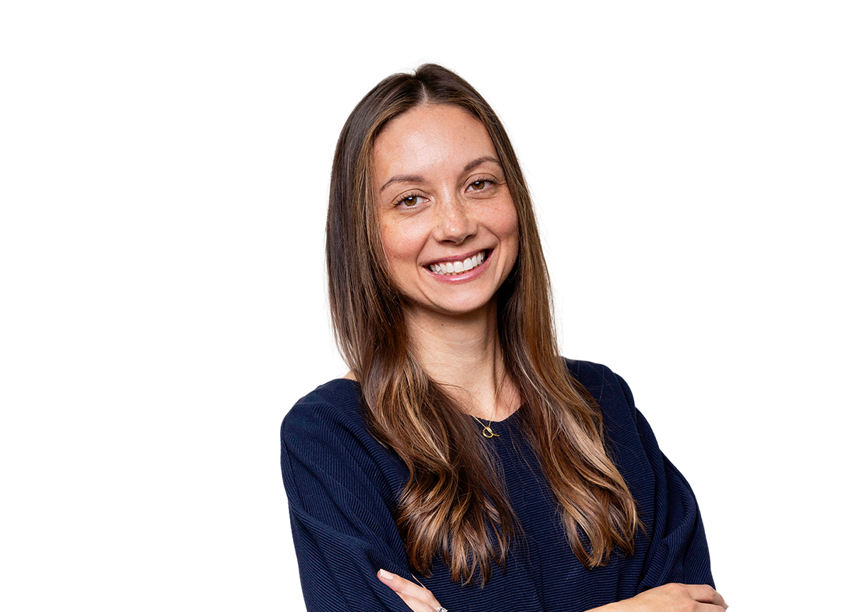Portrait of a smiling woman with long brown hair wearing a navy blue top and gold necklace, standing with arms crossed.