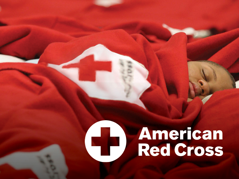 A young child sleeping wrapped in a red blanket with the American Red Cross logo, lying on a red bed or surface.