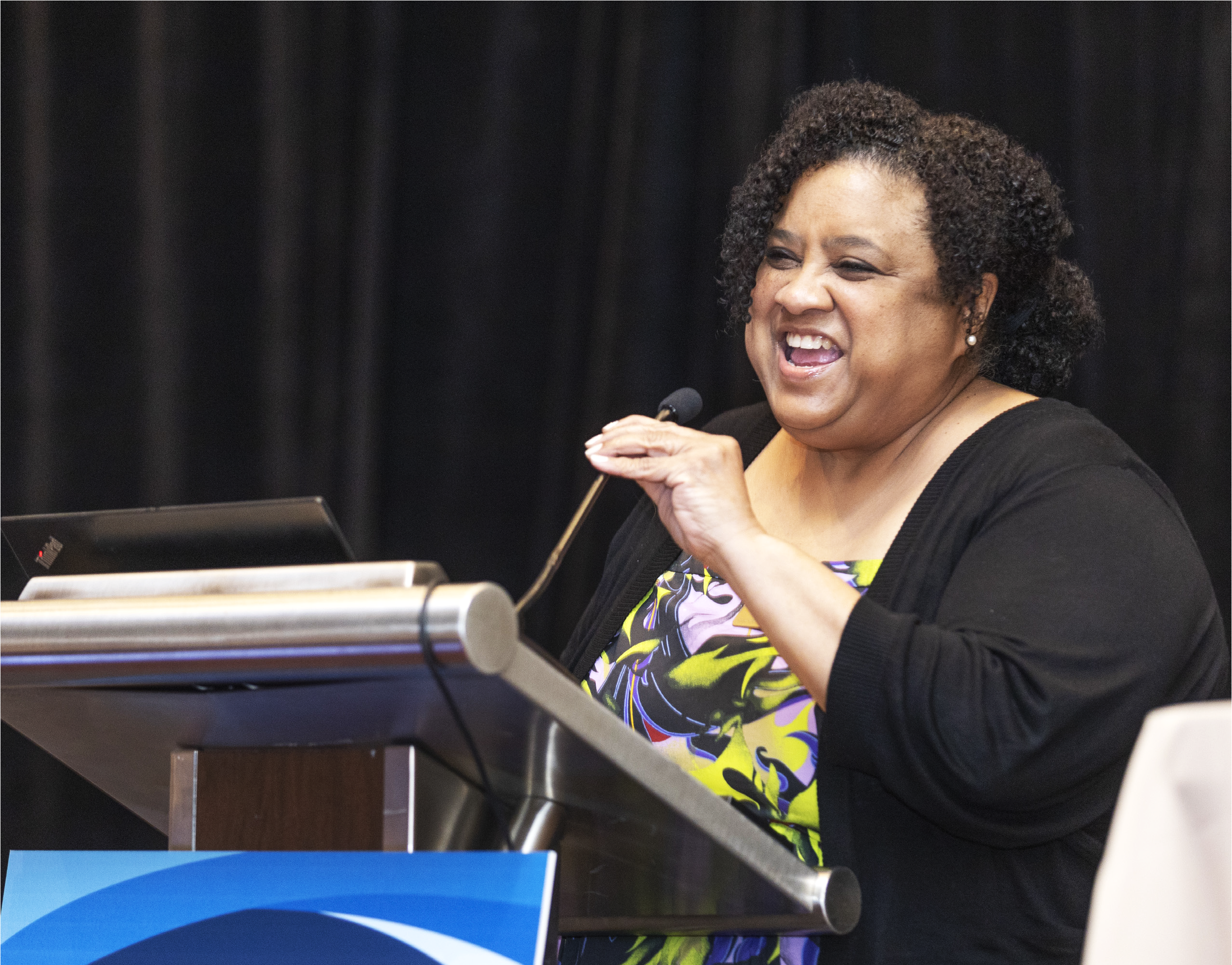 Woman with curly hair smiling and speaking at a microphone, seated at a podium, with a black backdrop behind her.