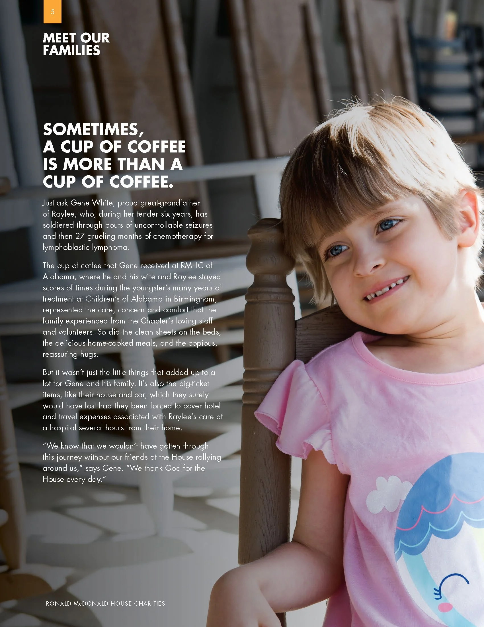 A young girl with blonde hair and blue eyes wearing a pink shirt with a rainbow unicorn graphic, smiling and leaning against a wooden bedpost in a brightly lit room.