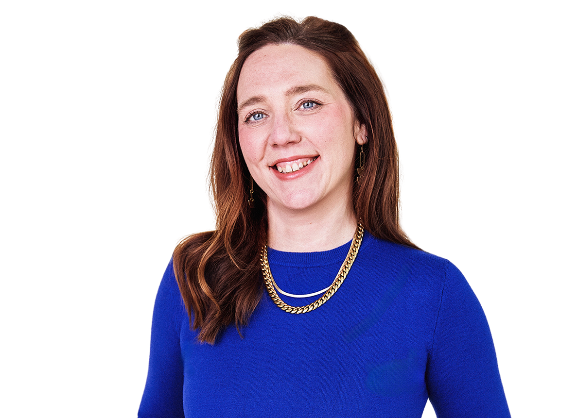 A woman with long brown hair, smiling, wearing a blue top, layered gold necklaces, and earrings against a multicolored background.
