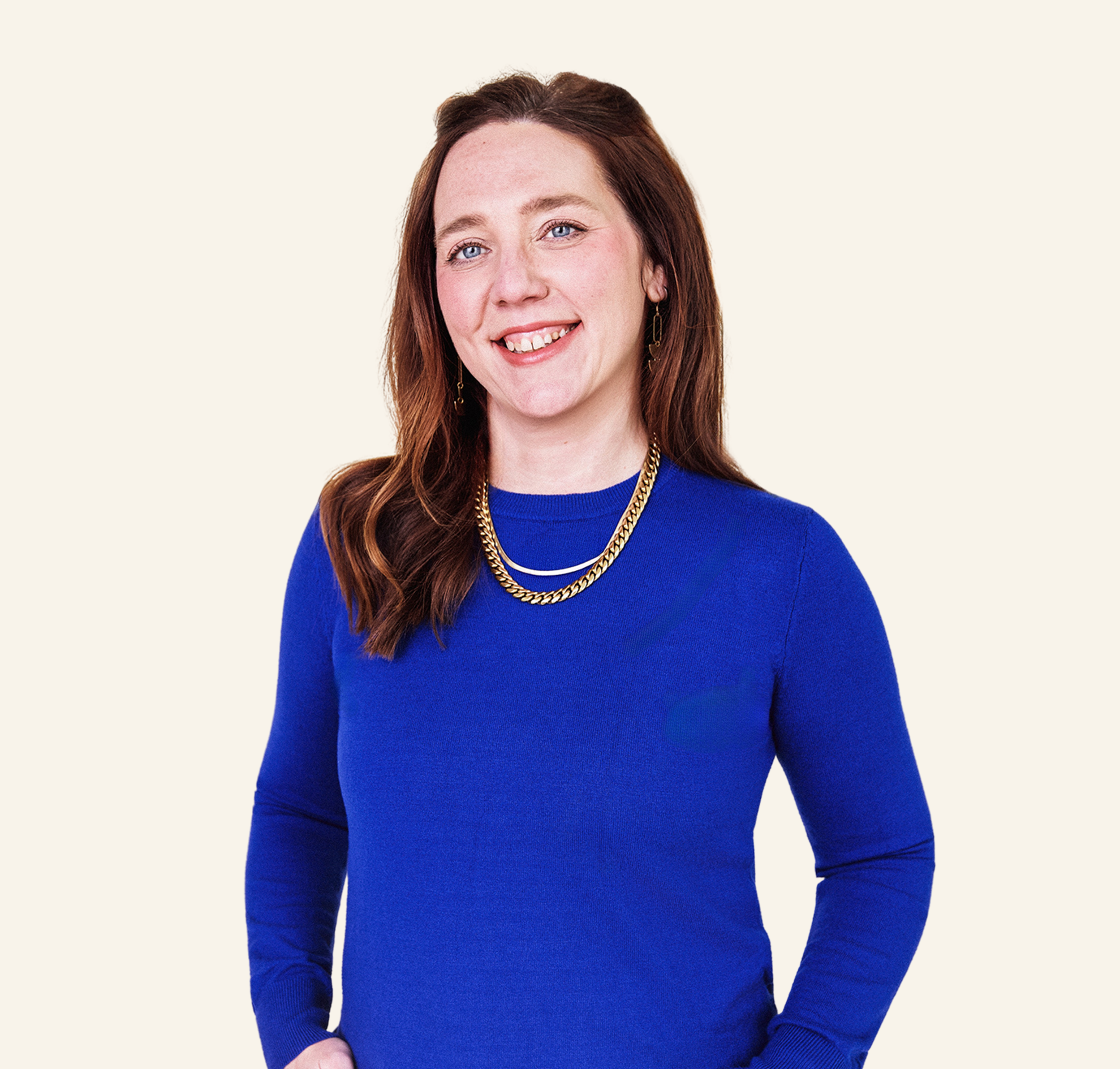 Portrait of a woman with long red hair wearing a royal blue top, gold chain necklaces, and earrings, standing against a plain light background.
