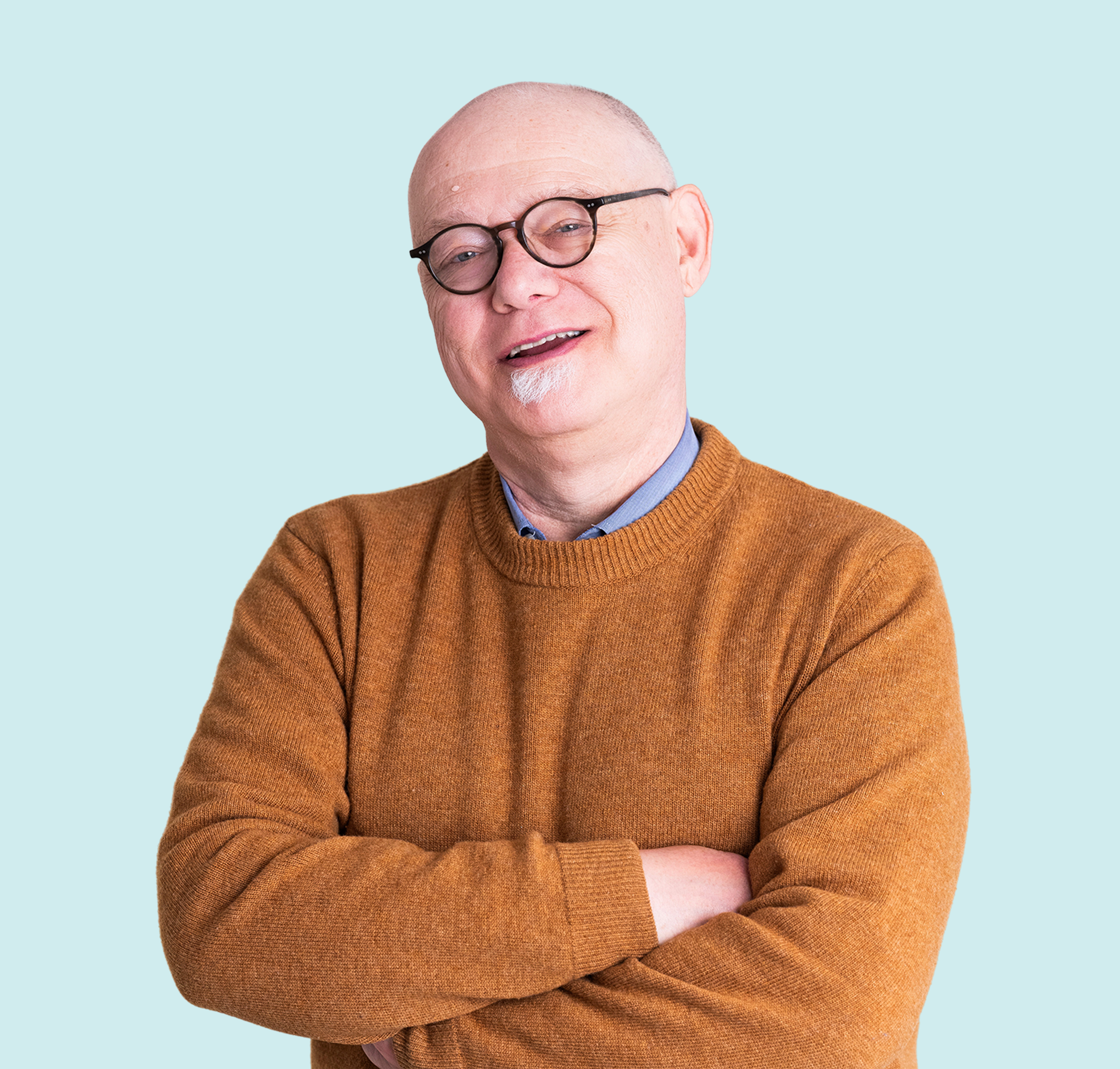 A middle-aged man with a bald head, glasses, and a goatee, wearing a brown sweater, standing with arms crossed against a light blue background.
