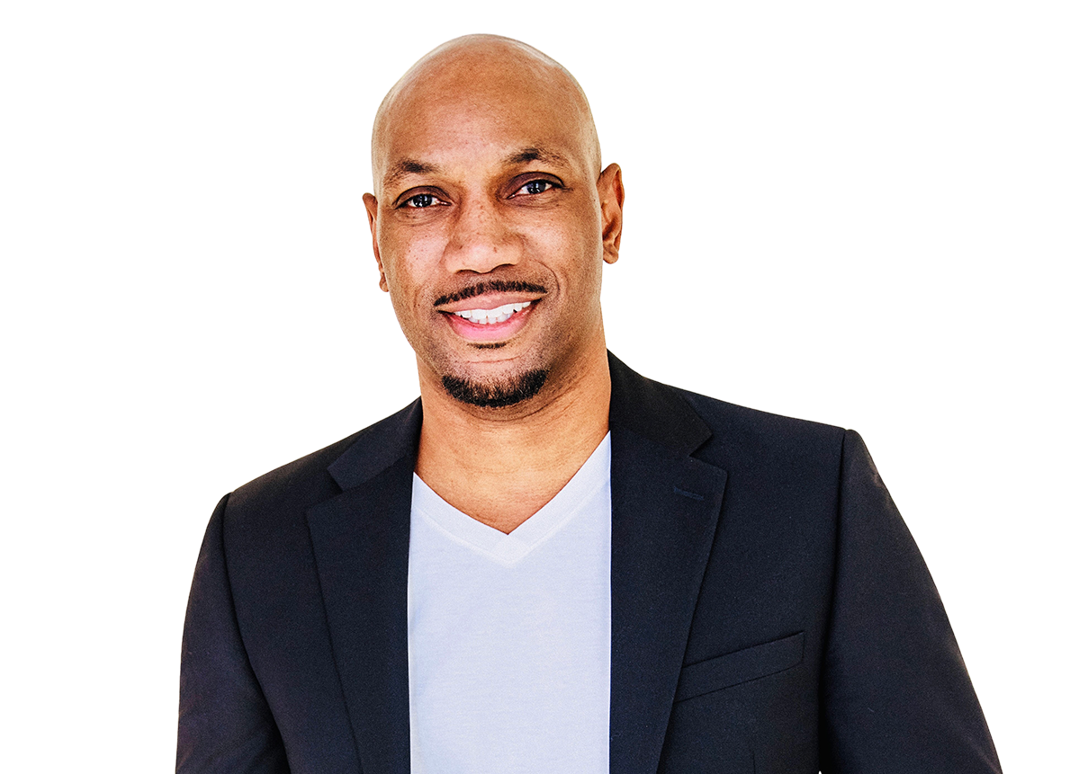 Man with a shaved head and mustache, wearing a black blazer and white V-neck shirt, smiling