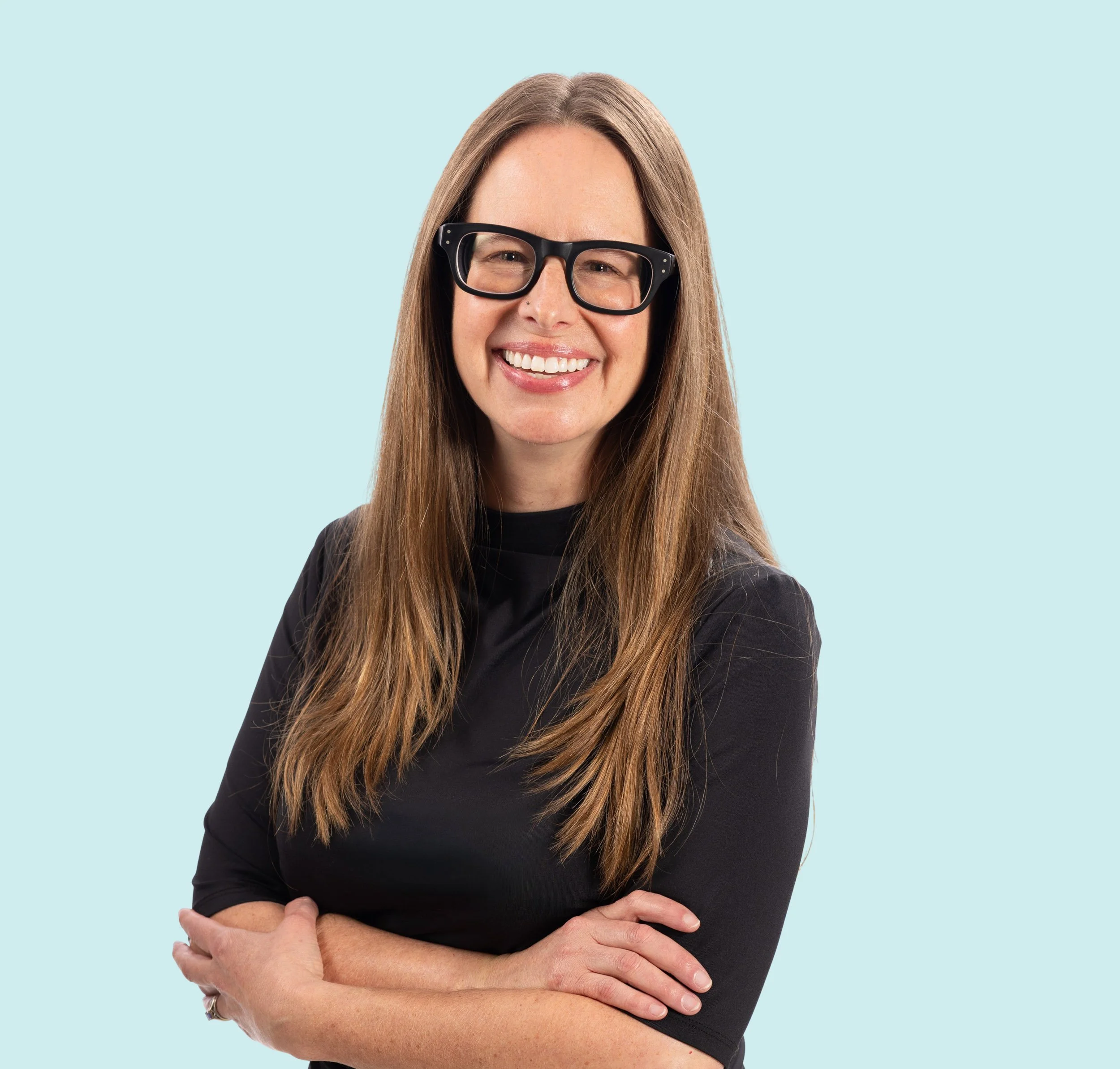 Portrait of a cheerful woman with long hair wearing large black glasses, crossing her arms, against a light blue background.