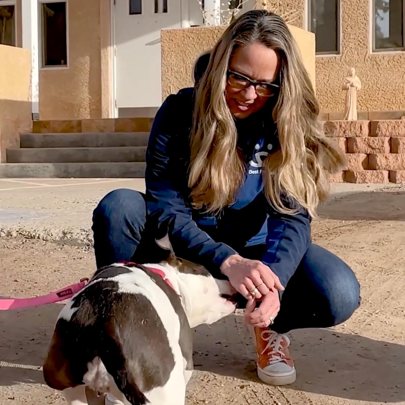 A woman with long blonde hair and glasses crouching on the ground, petting a small black and white dog with a pink leash outside a beige house with steps and windows.
