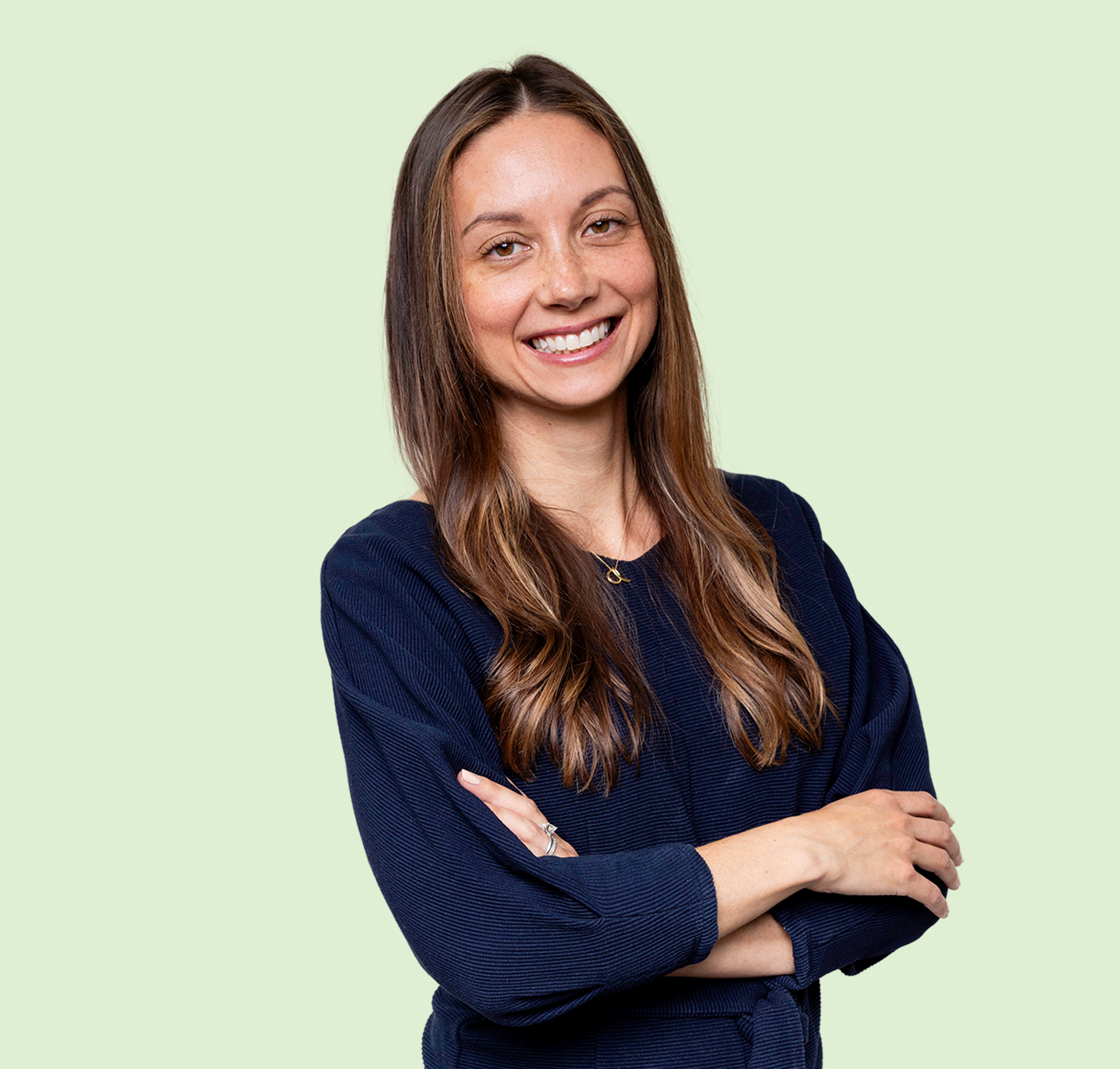 Smiling woman with long brown hair wearing a dark blue top, standing against a light green background.