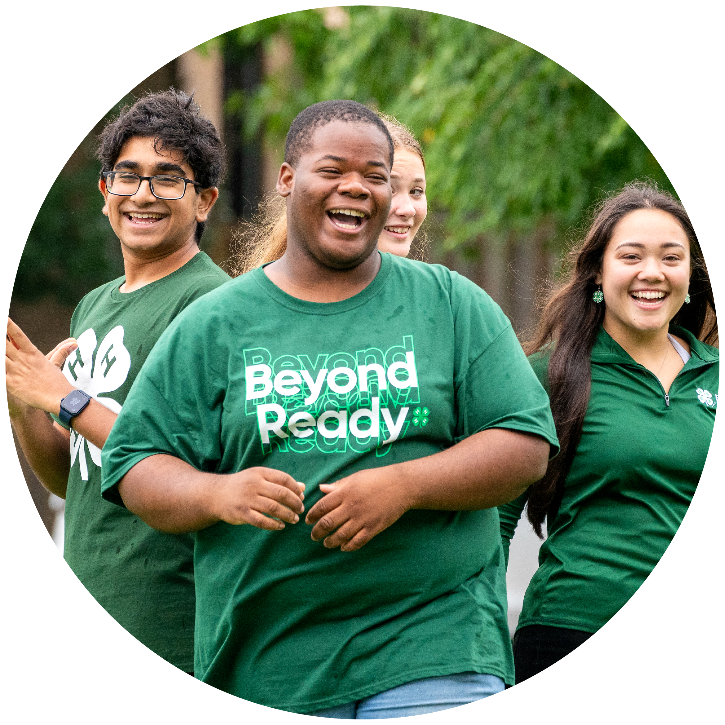 A group of four diverse smiling young people wearing green t-shirts with the words 'Beyond Ready' printed on them, standing outdoors with a blurred green background. They are laughing and appear happy.