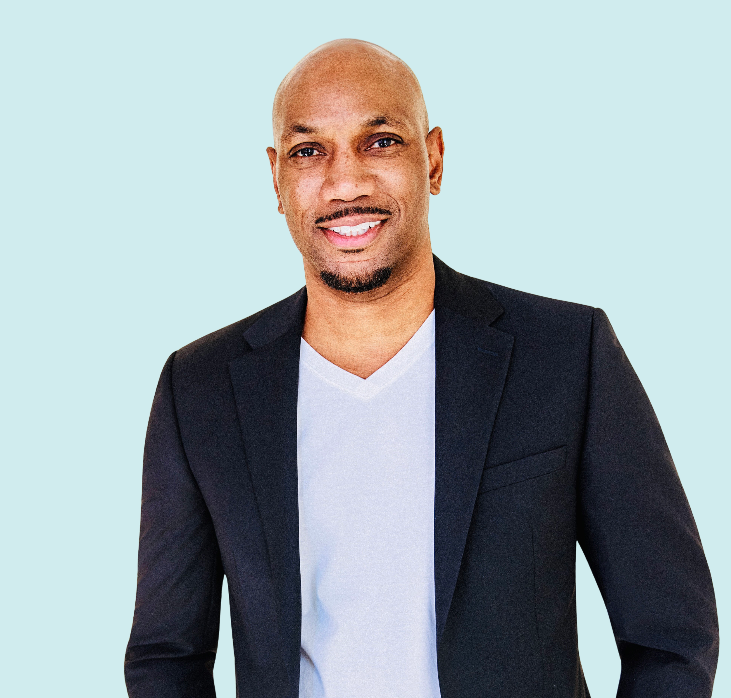 Headshot of an African American man with a shaved head, goatee, wearing a black blazer over a white V-neck shirt, smiling against a light blue background.