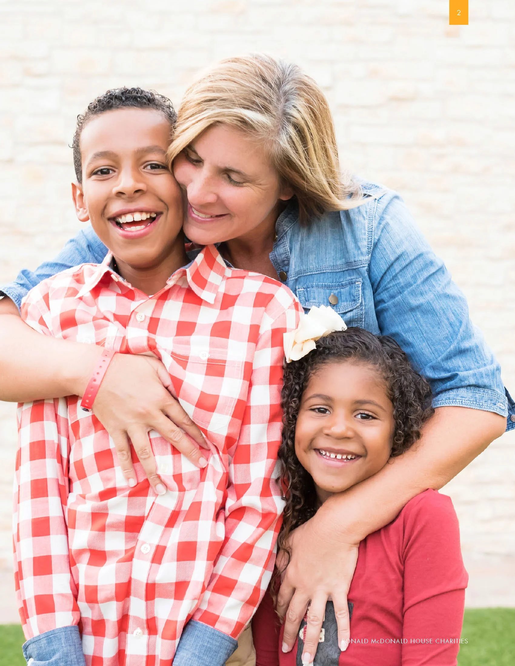 Happy woman and two young girls hugging outdoors by a beige brick wall. The woman has blonde hair and is wearing a blue denim shirt. The girl is smiling, wearing a red and white checkered shirt, and has curly hair. The other girl, partially visible, has dark curly hair and a flower hair clip.