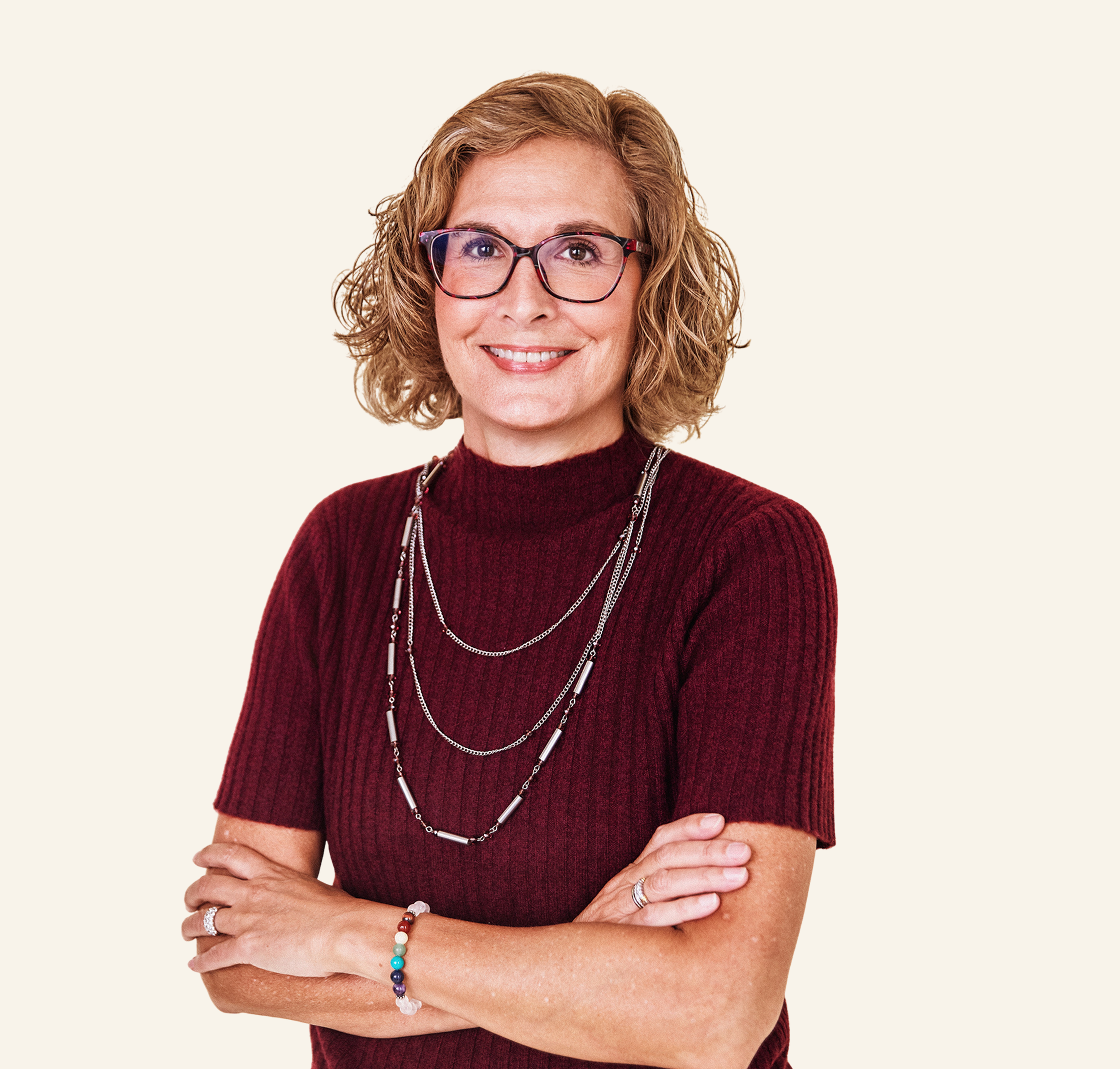 A woman with curly hair wearing glasses and a maroon sweater, standing with arms crossed against a plain light background.