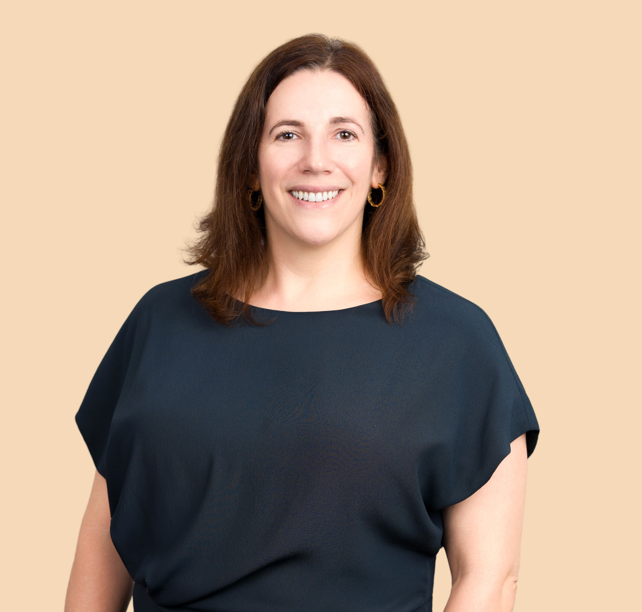 Close-up of woman smiling, wearing a dark top, with earrings and brown hair, against beige background.