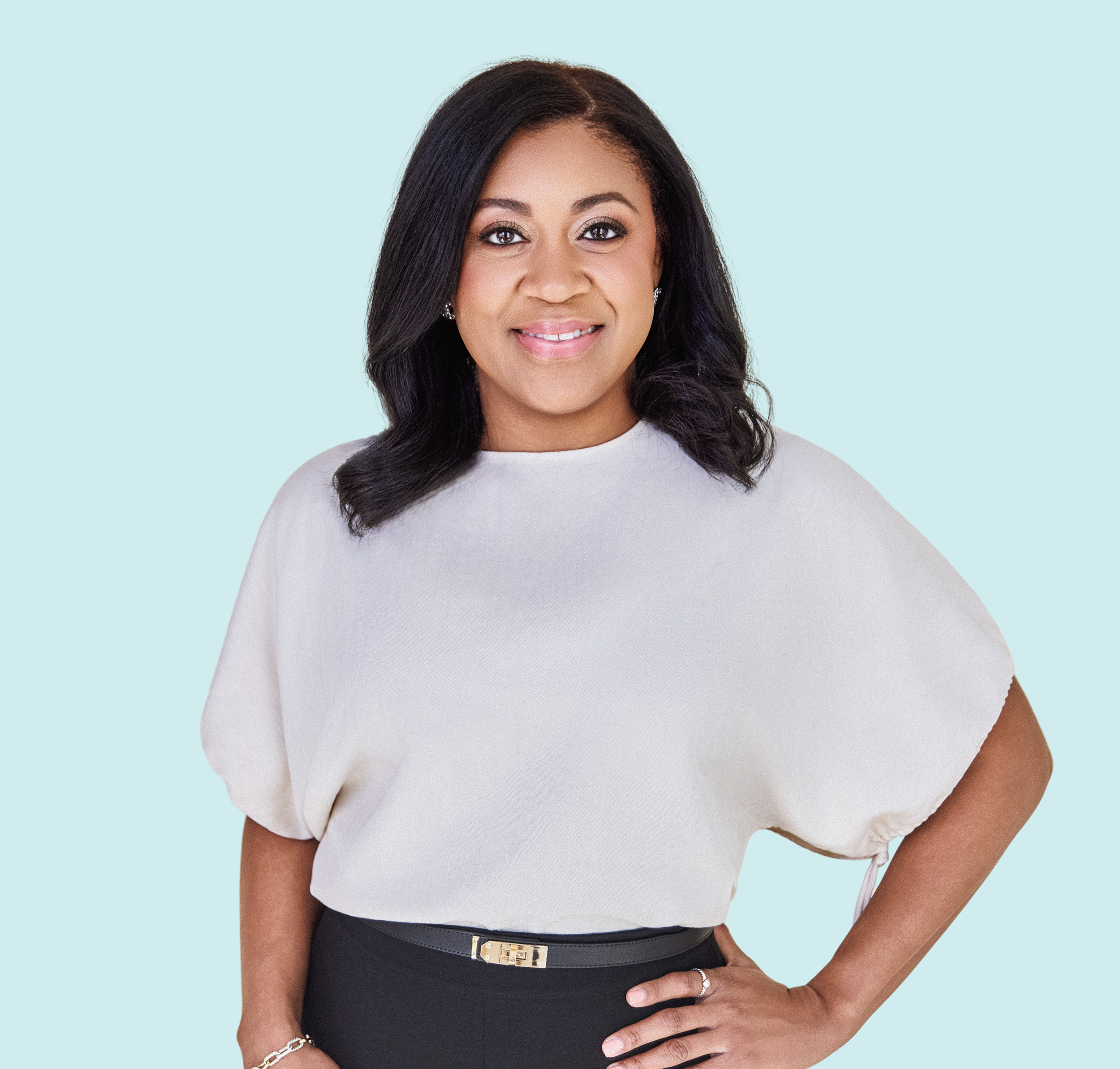 A woman with dark hair styled in loose waves, wearing a white top with puffed sleeves and a black skirt, standing against a light blue background.