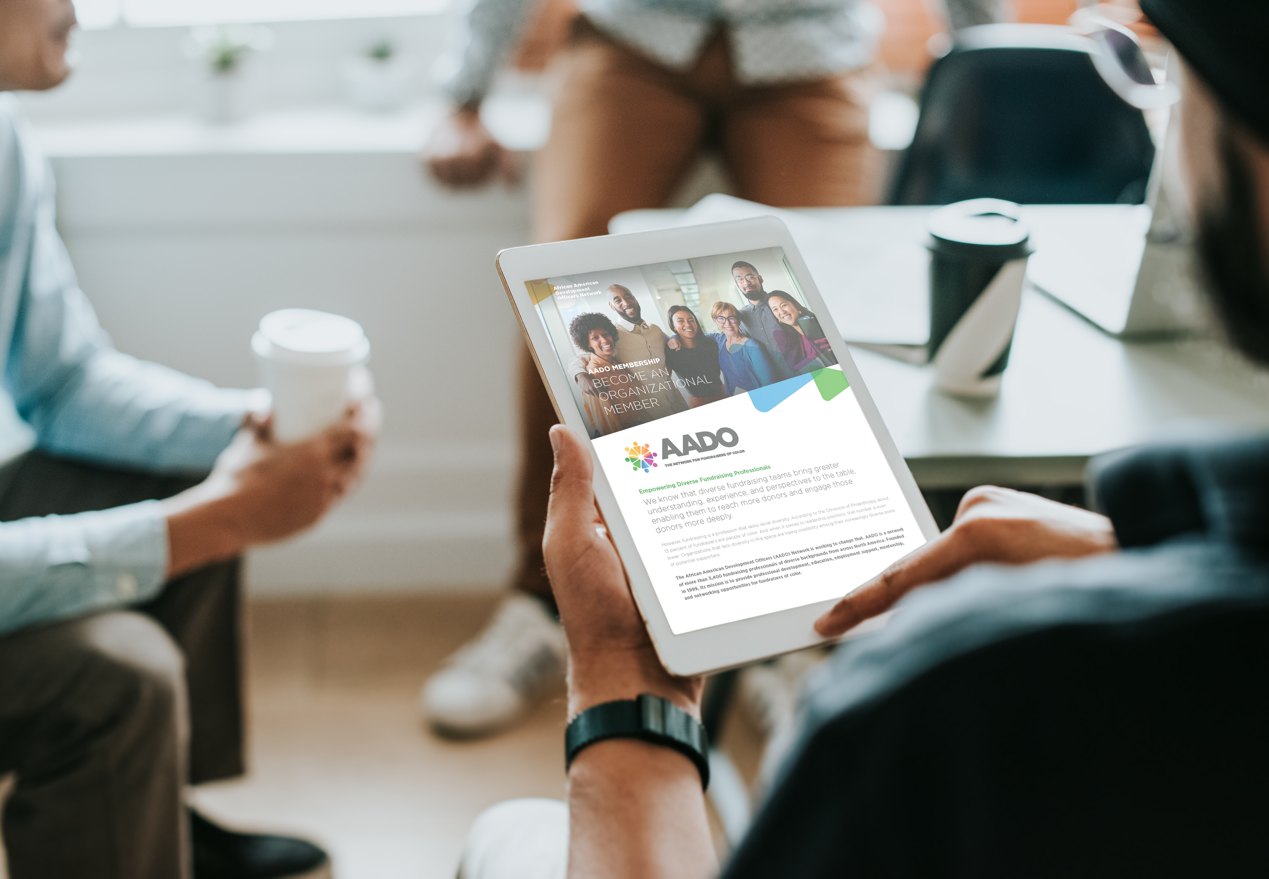 Person holding a tablet displaying the AAIDO organization website with a group photo of diverse smiling people, during a meeting.