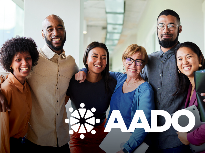 Group of six diverse smiling people standing together in an office, with an AADO logo at the bottom.