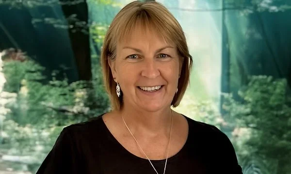 A smiling middle-aged woman with short blonde hair, wearing earrings and a black top, standing outdoors with a green, wooded background and glass windows.