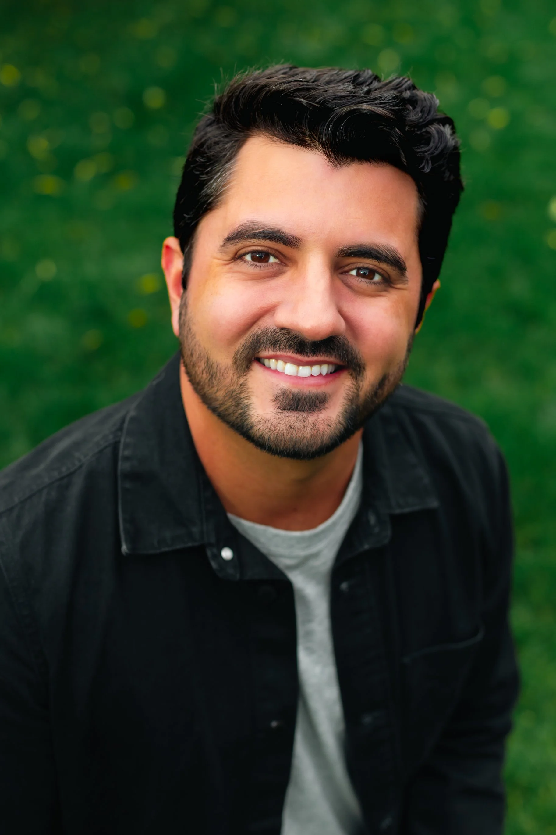 Smiling man with dark wavy hair and a short beard wearing a black button-up shirt over a gray t-shirt, photographed outdoors against a green grassy background in a close-up headshot.