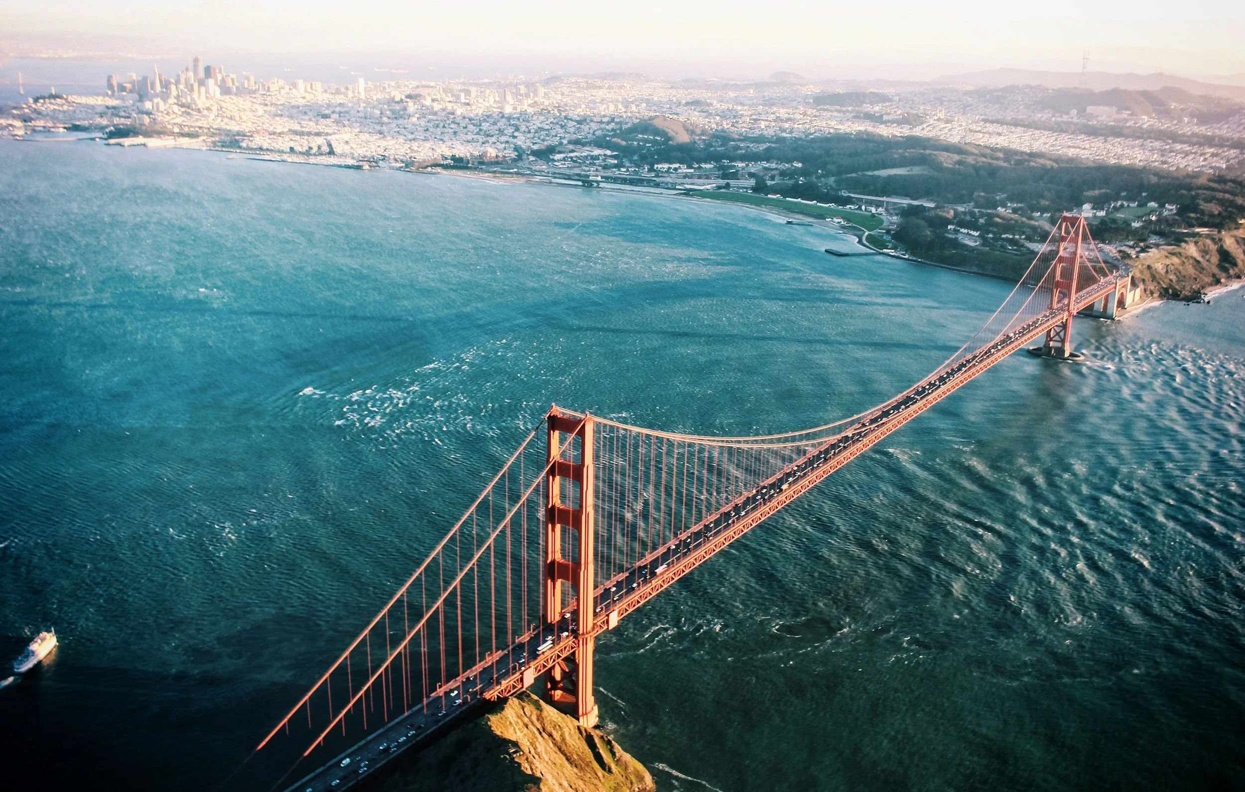 Aerial view of the Golden Gate Bridge over water with city skyline and hills in the background during daylight.
