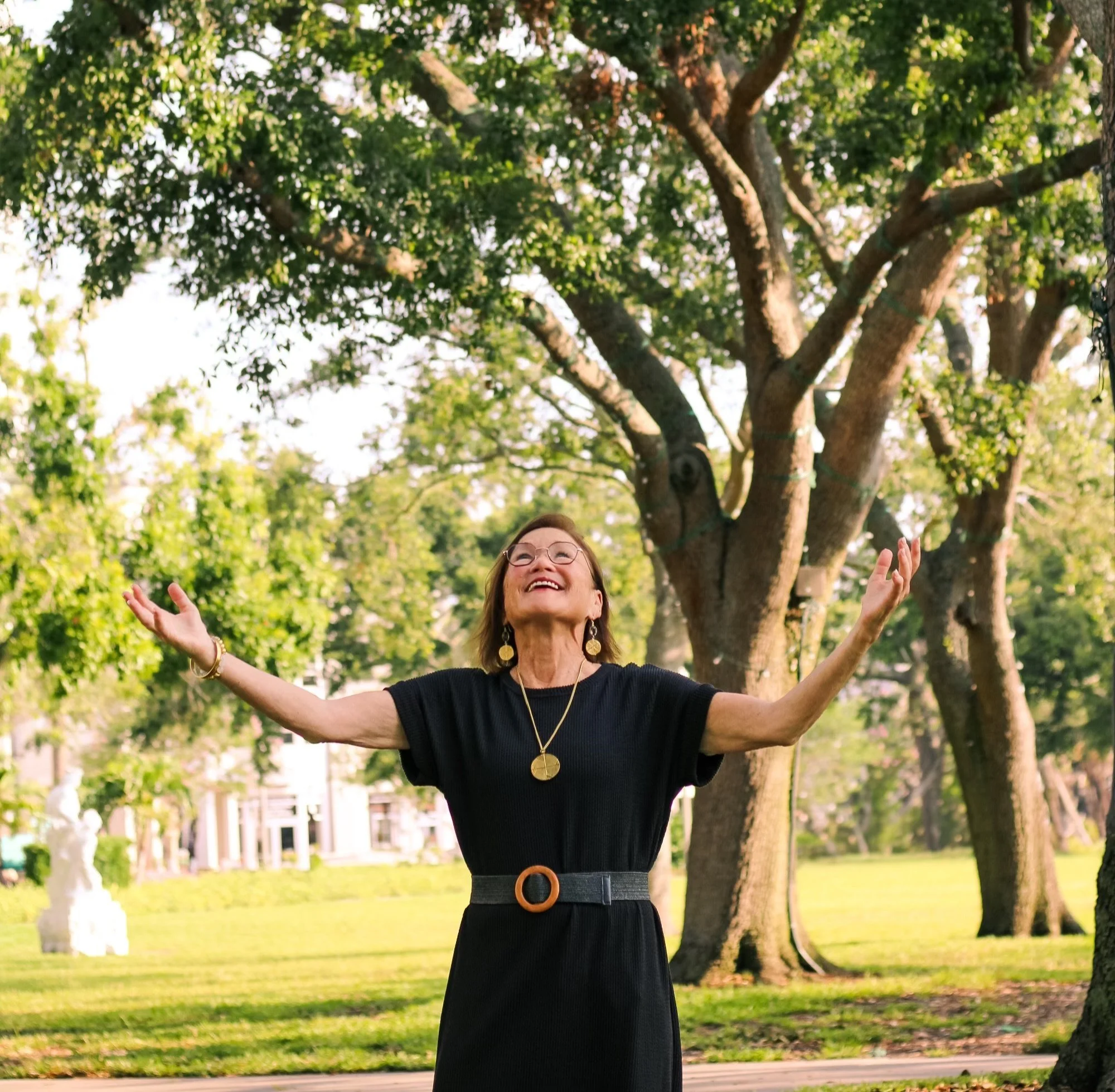 A woman smiling with arms outstretched in a park with green trees and grass.