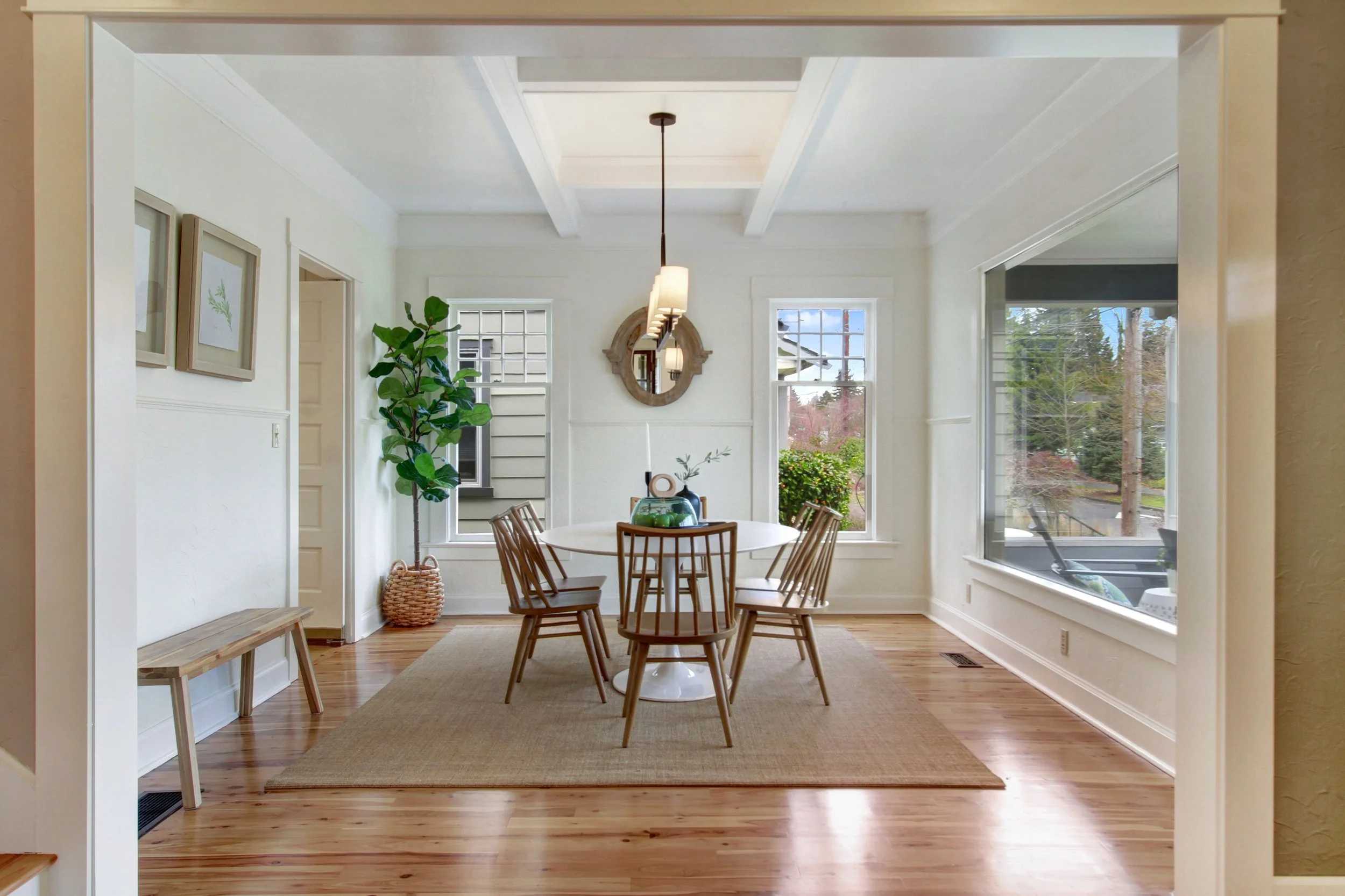 A bright dining room with a round table and six wooden chairs. Large windows let in natural light, and there’s a view of trees outside. A potted plant is near the doorway, and a beige rug covers part of the hardwood floor.