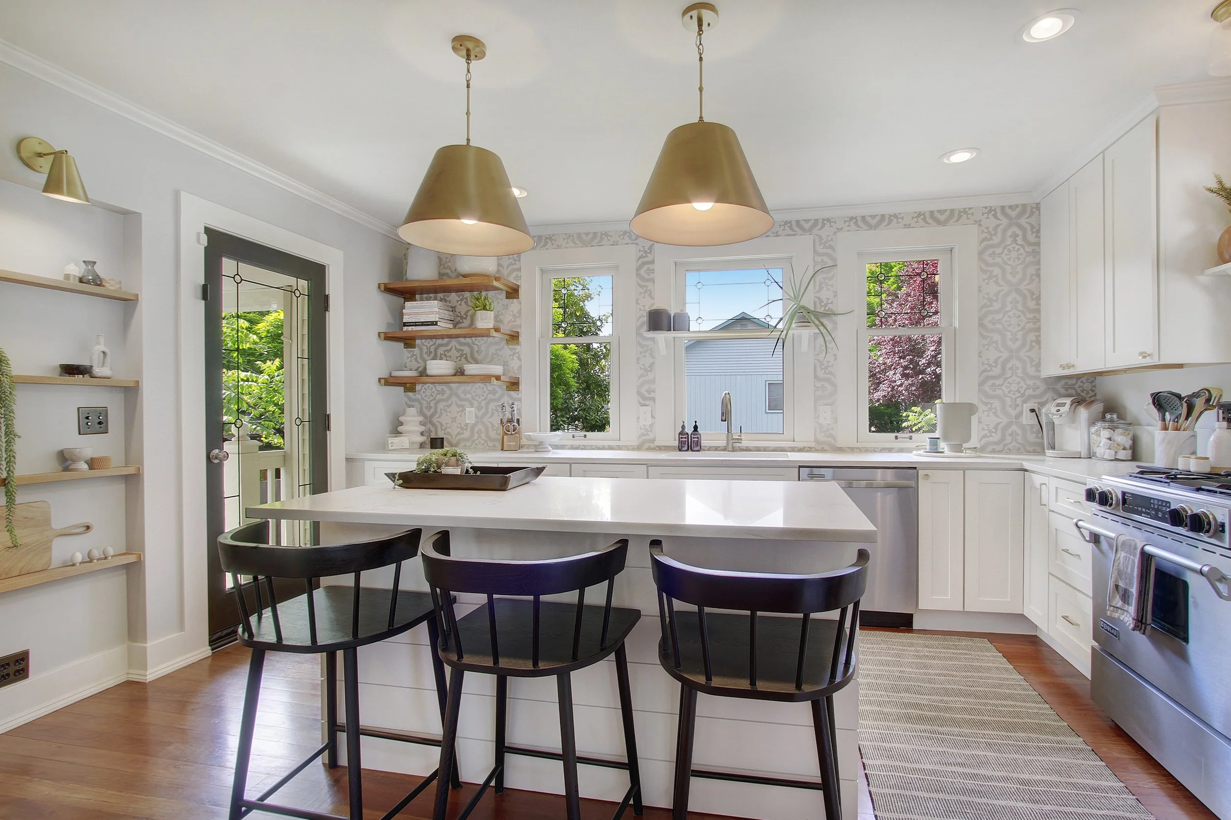 Bright kitchen with white cabinets and a center island, three black chairs around the island, wooden shelves on the wall with decorative items, large windows showing green trees outside, pendant lights hanging from the ceiling, and a stove in the corner.