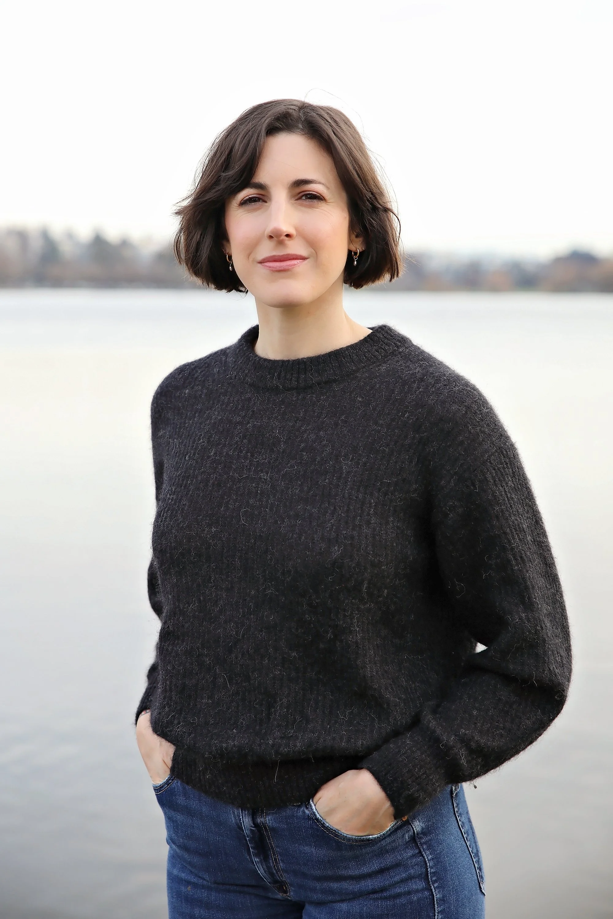 A young woman with short dark hair, wearing a black sweater and blue jeans, standing outdoors near water with a blurred natural background.